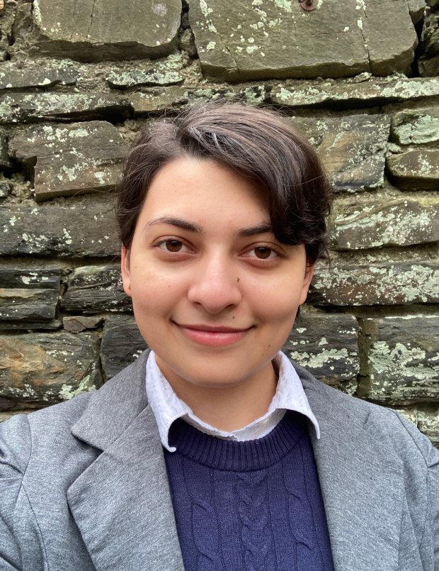 Photograph of a person in a gray blazer and a dark blue sweater over a collared shirt. Short brown hair and light brown skin. The background is a lichen-mottled stone wall.